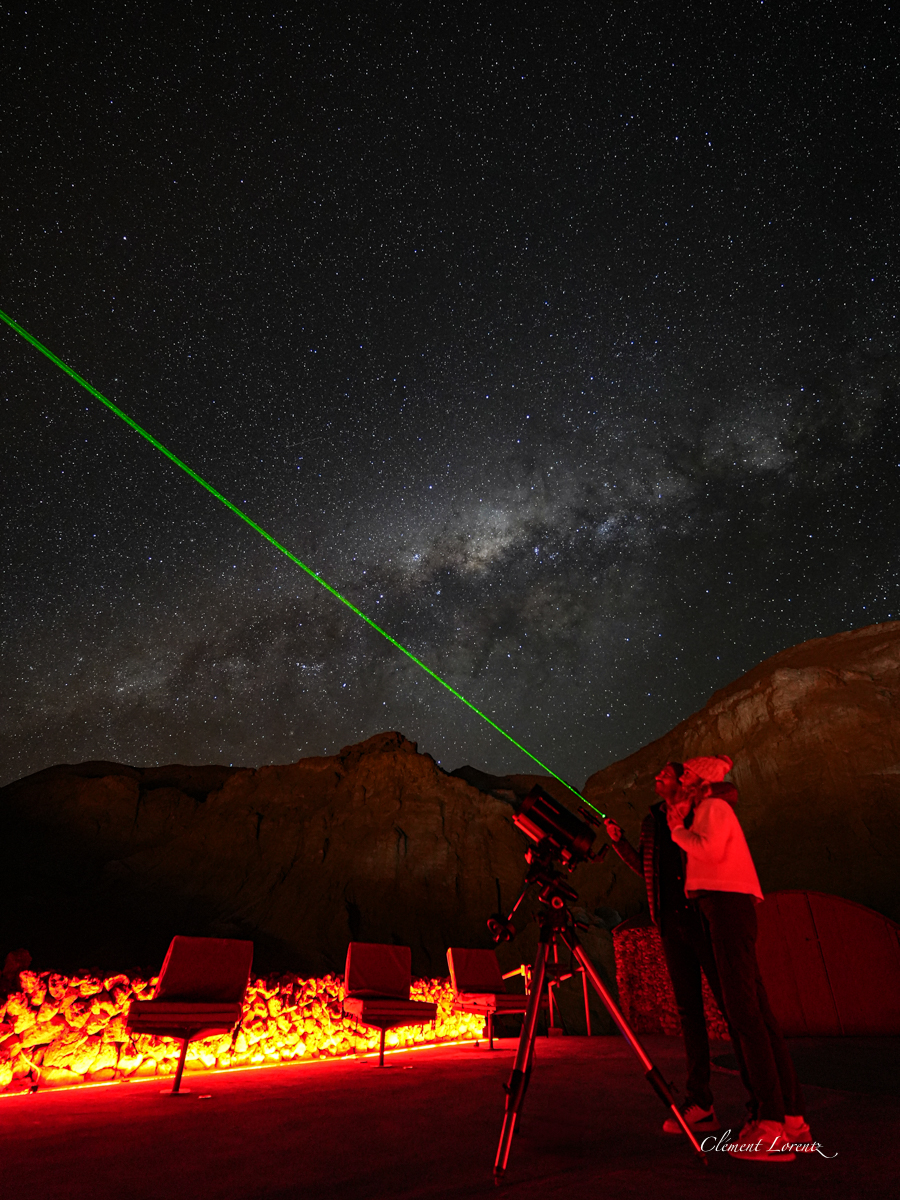 Astro photo Clement Lorentz Astrophotography scene in the Atacama Desert, Chile, showing a couple next to a telescope as the man points a green laser toward the starry night sky.